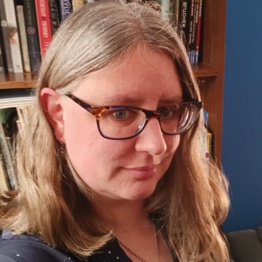 A middle aged white women with greying hair and glasses looking at the camera slightly smiling with a bookshelf in the backgound